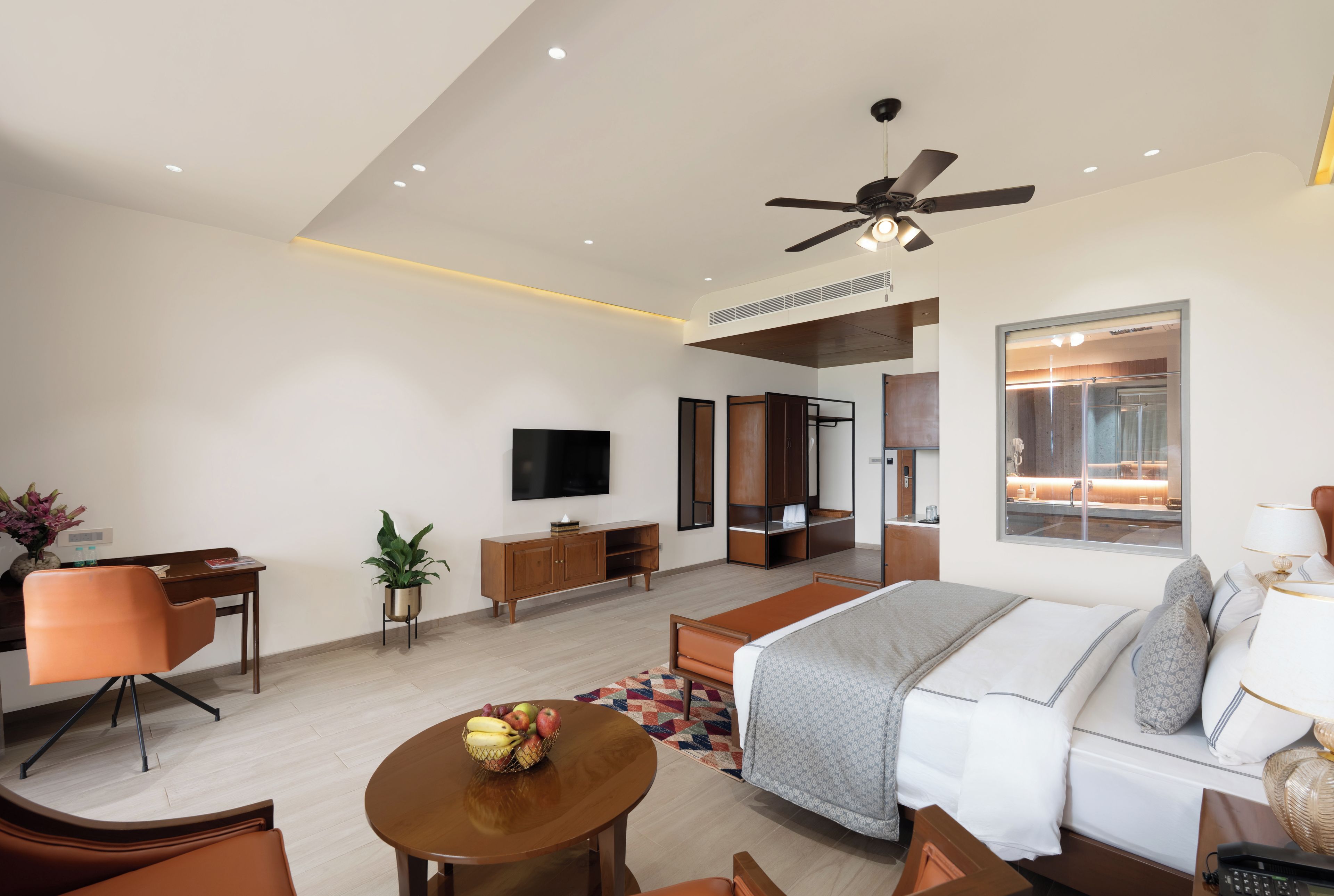 A modern hotel room with a bed, seating area, TV, and a bathroom visible through a glass door.