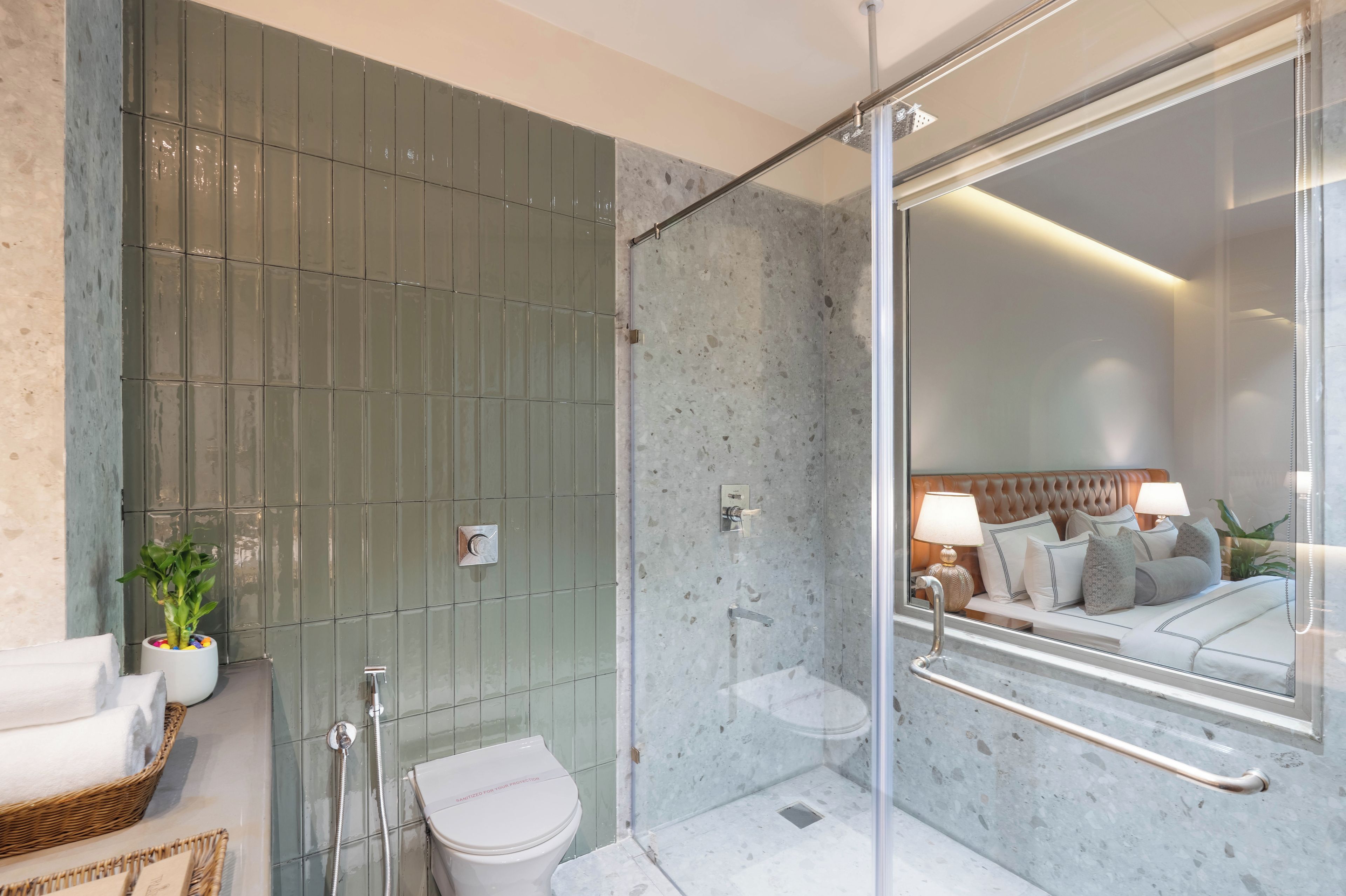 Et moderne badeværelse med en glasbrusekabine, toilet og en synlig seng med et natbord i det tilstødende rum.