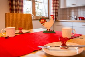 Uma mesa de jantar com um jogo americano vermelho, duas canecas brancas, um copo vermelho, um prato branco e uma decoração de galo com detalhes em vermelho e verde.