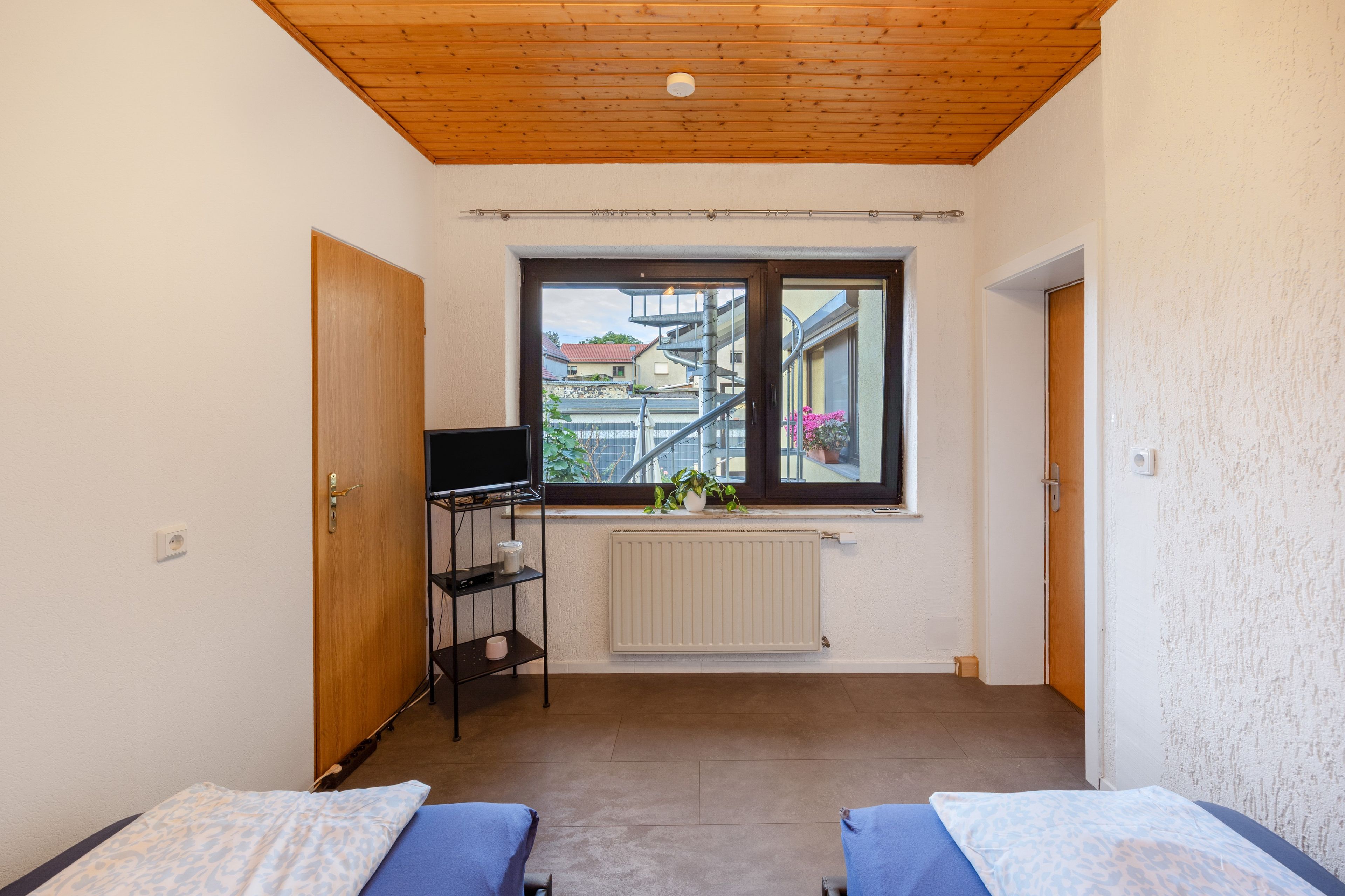 A room with two beds, a window with a view of buildings, a radiator, and a shelf with items.