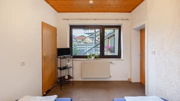 A room with two beds, a window with a view of buildings, a radiator, and a shelf with items.