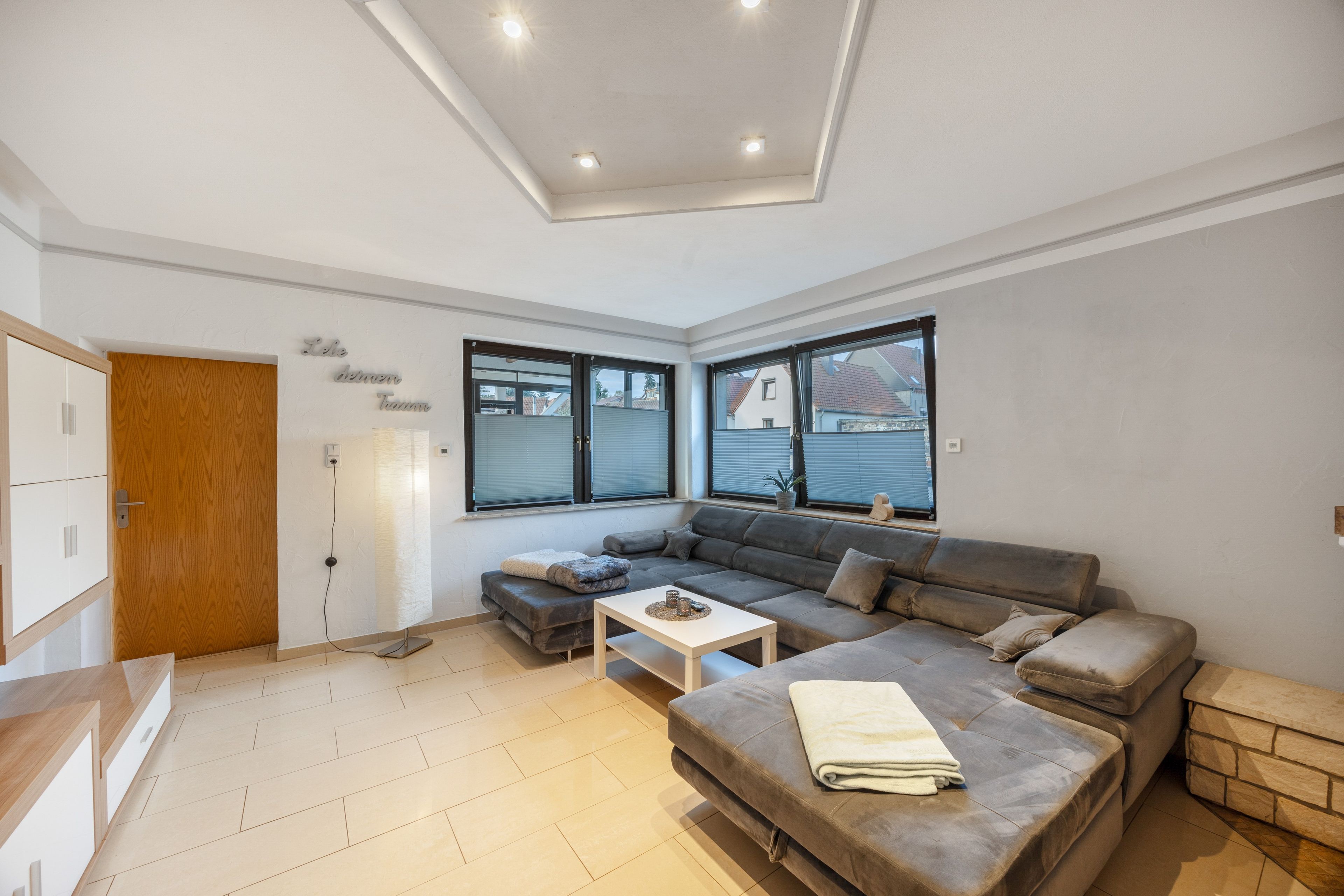 A modern living room with a sofa, a coffee table, and a dining area. There is a wooden door and a window with blinds.