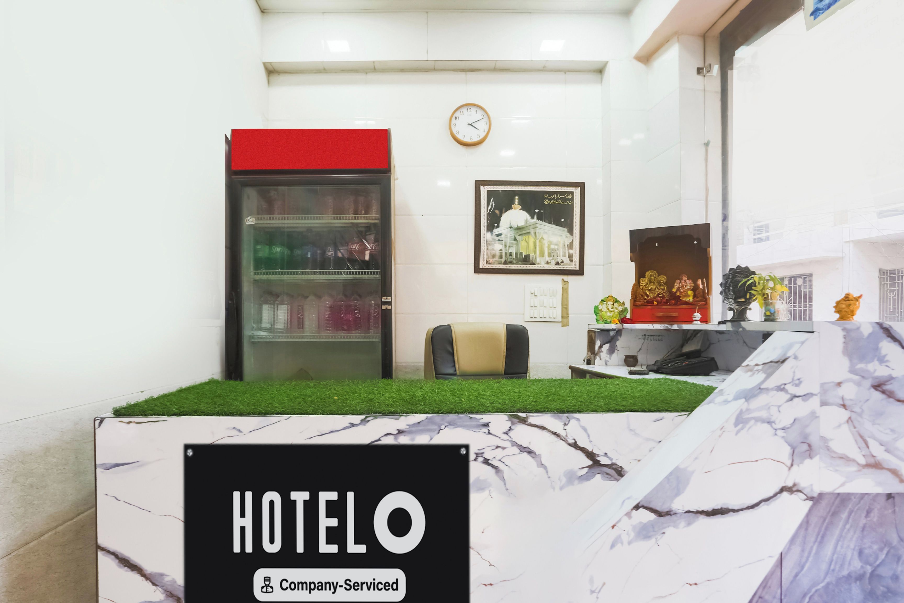 Zona de recepción de un hotel con mostrador, un reloj, un refrigerador y un cuadro en la pared.