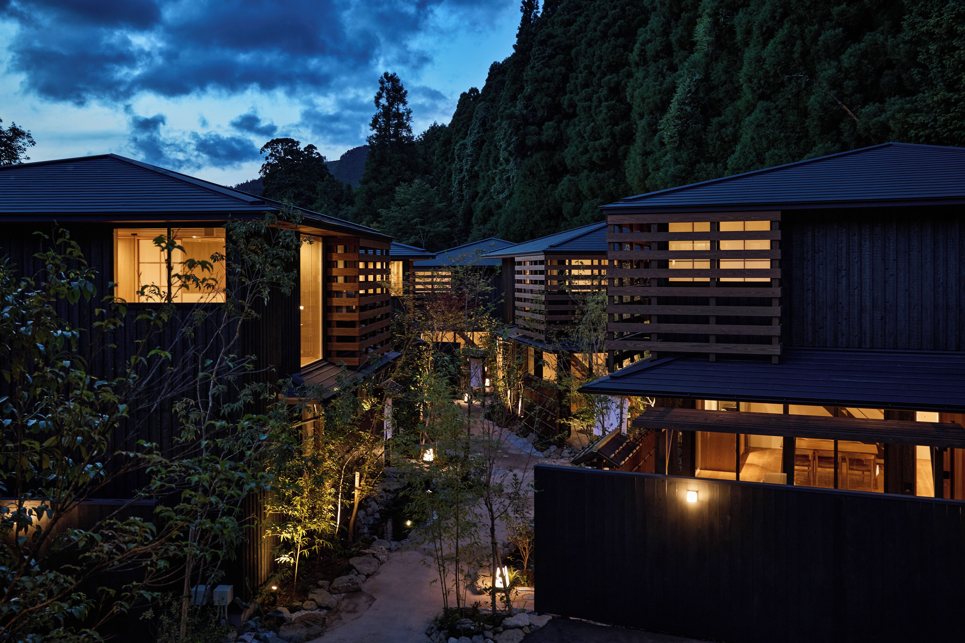A row of traditional wooden buildings with large windows, surrounded by a garden with rocks and plants, under a twilight sky.