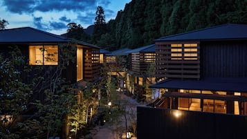 A row of traditional wooden buildings with large windows, surrounded by a garden with rocks and plants, under a twilight sky.