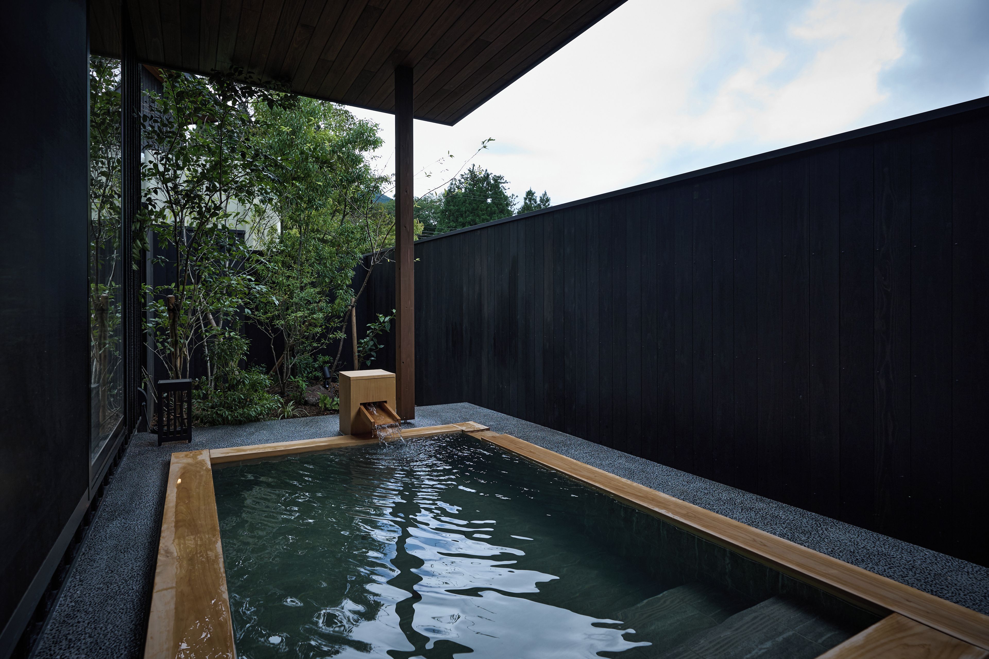 A rectangular pool with a wooden edge, surrounded by a dark fence, and a small wooden stool near the water's edge.