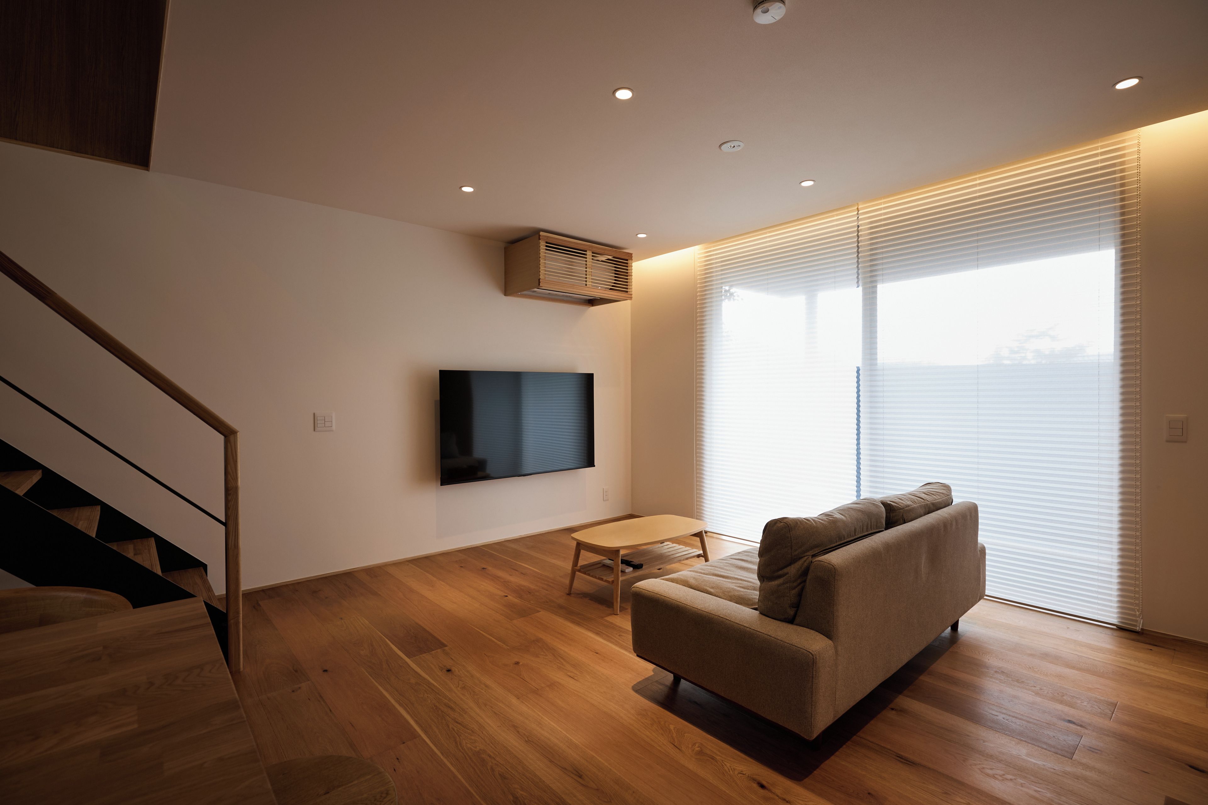 A modern living room with a sofa, a flat-screen TV mounted on the wall, a small table, and a staircase leading to an upper level.