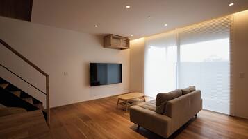 A modern living room with a sofa, a flat-screen TV mounted on the wall, a small table, and a staircase leading to an upper level.