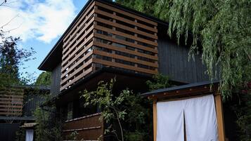 A traditional Japanese-style building with a sloped roof, wooden lattice structure, and a view of a garden with trees and a hanging plant.