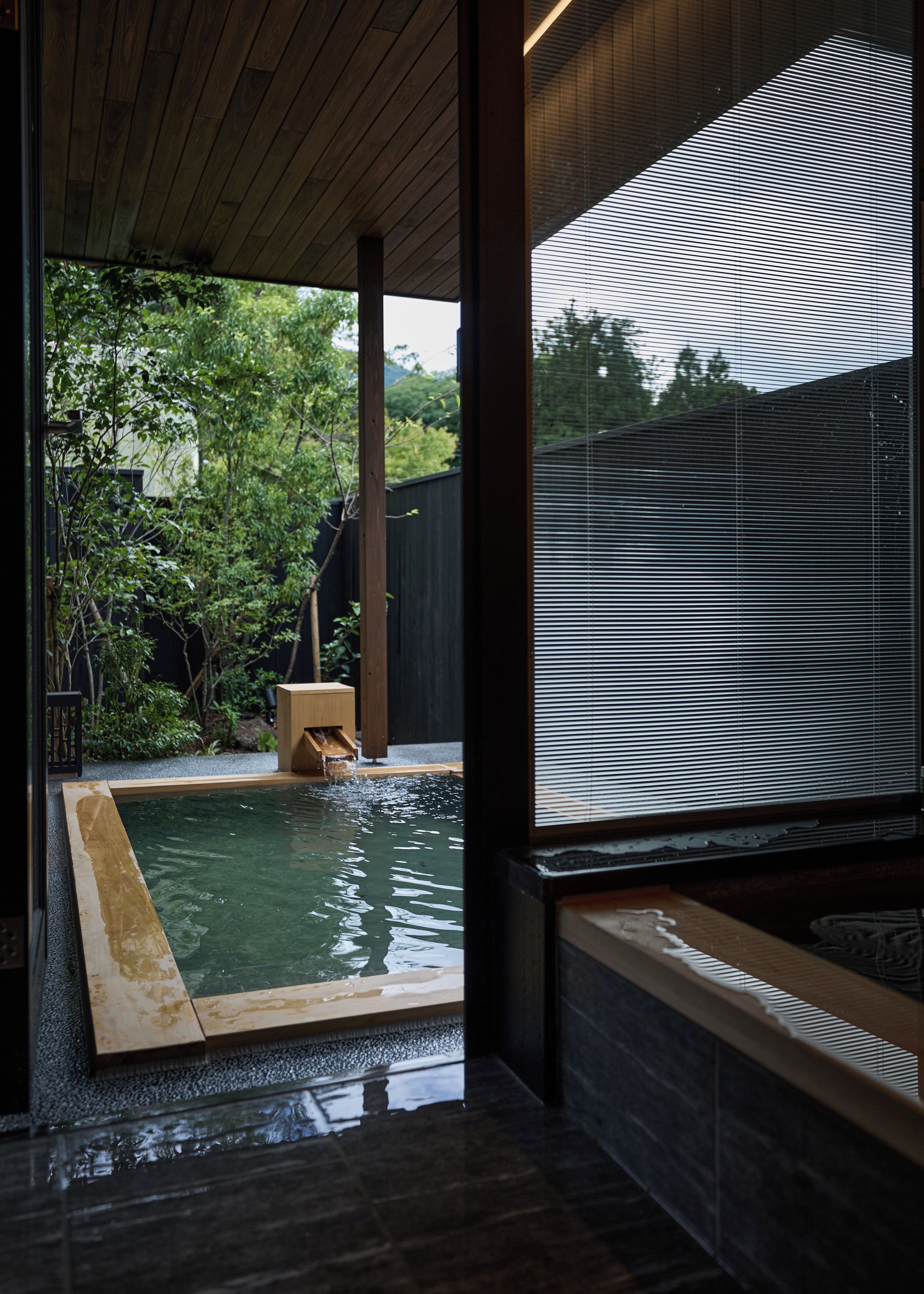 Una habitación tradicional japonesa con techo de madera, suelo de esteras de tatami, un pequeño banco de madera y una ventana con vistas a un jardín.