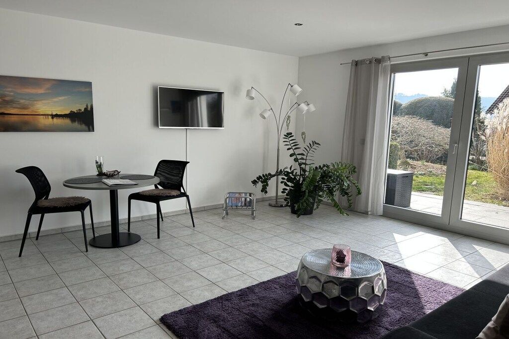 Ein modernes Wohnzimmer mit Essbereich, einem an der Wand befestigten Flachbildfernseher und einem großen Fenster mit Blick ins Freie.