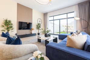 Una sala de estar moderna con televisor de pantalla plana, sofás, una mesa de centro y ventanas amplias con vistas al exterior.