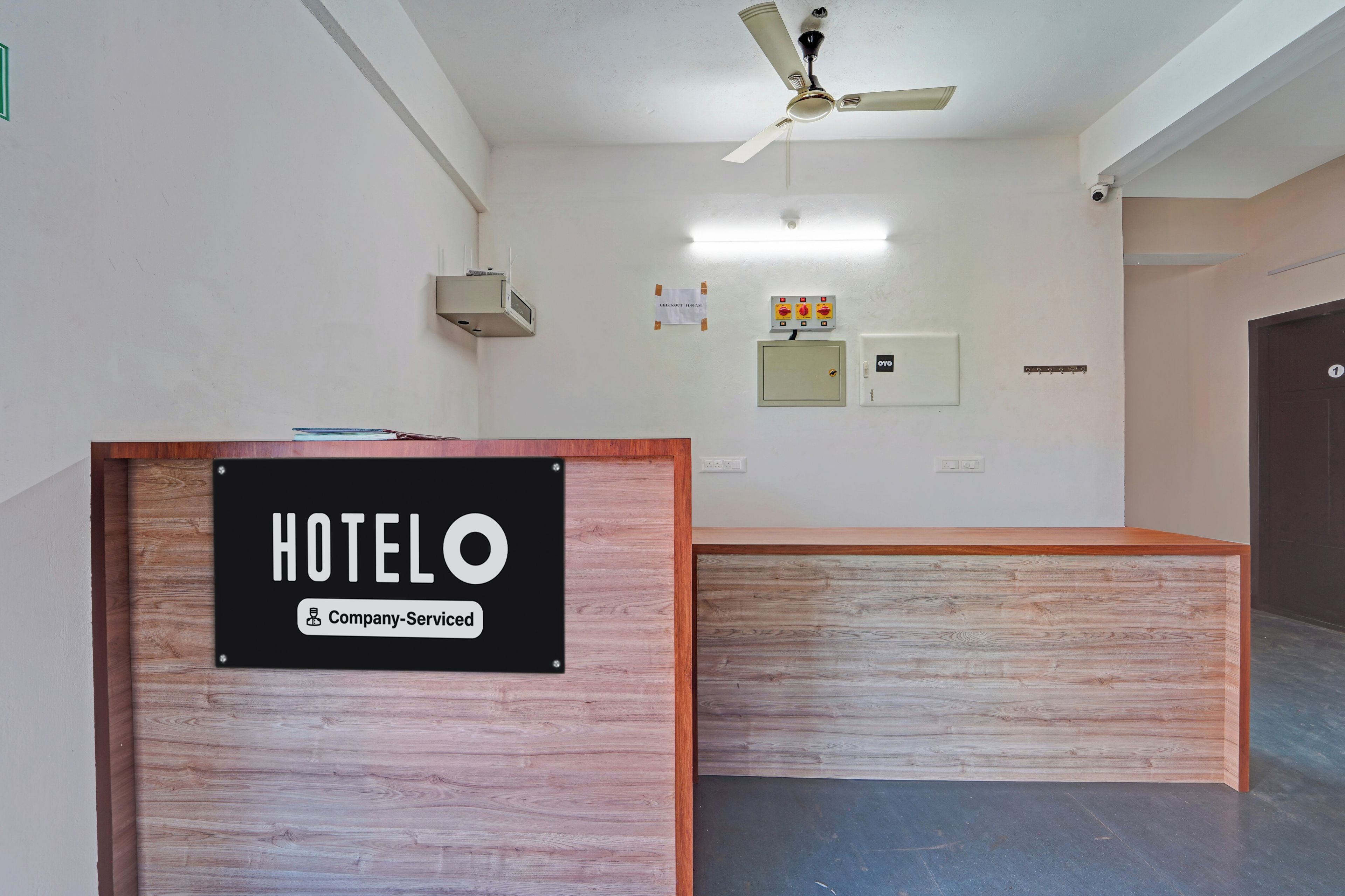 A hotel reception desk with a sign indicating it is company-serviced. There is a ceiling fan and a wall-mounted light fixture above the desk.