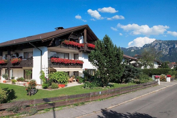 Un edificio de dos pisos con balcones, una cerca de madera y montañas al fondo.