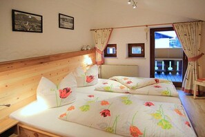 Un dormitorio con cabecera de madera, dos camas con ropa de cama floral, una mesita de noche, un ventanal con cortinas y vistas a un balcón.