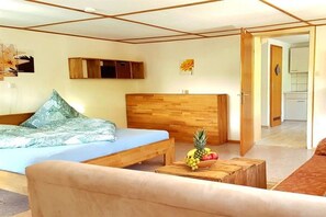 Quarto com cama, cabeceira de madeira, uma mesinha com frutas e uma porta que dá para uma cozinha.