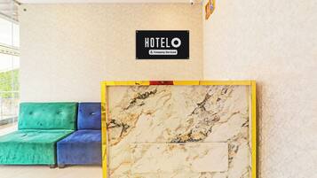 Zona de recepción de un hotel con pared de mármol, cartel que indica que es un hotel de servicios corporativos y una sala de espera con sofás tapizados en tonos verde y azul.
