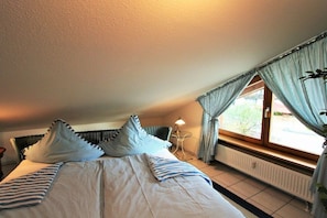 Quarto com cama, almofadas, mesa de cabeceira, luminária e janela com cortinas.