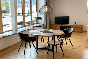 Uma sala de estar moderna com uma mesa de jantar redonda, cadeiras pretas, televisão e uma grande janela com vista para o exterior.