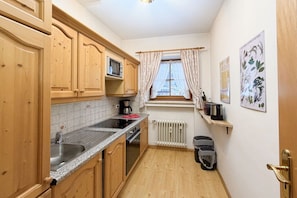 A compact kitchen with wooden cabinets, a microwave, a sink, and a window with curtains. - Holiday apartment with 1 bedroom (Garmisch-Partenkirchen)