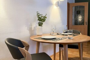 A dining area with a round wooden table set for two, a white vase with a plant, and a door with a glass panel. - Holiday apartment with 1 bedroom (Garmisch-Partenkirchen)
