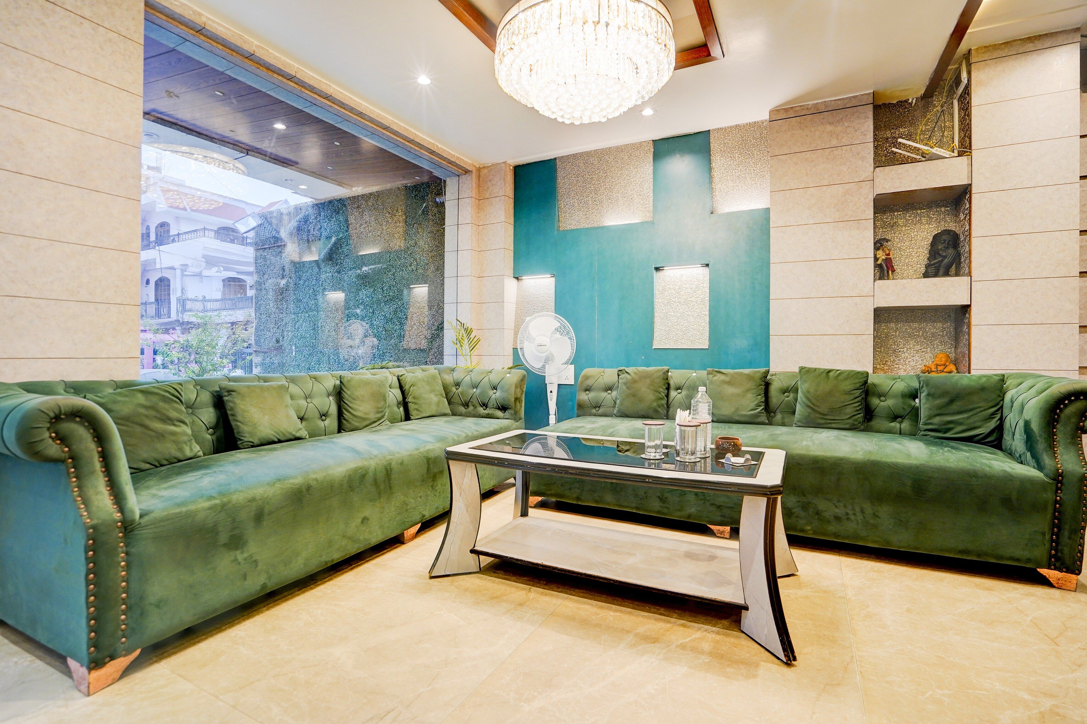 A green upholstered sofa with a glass coffee table, a chandelier, and a view of a building through a window.
