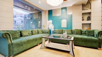 A green upholstered sofa with a glass coffee table, a chandelier, and a view of a building through a window.