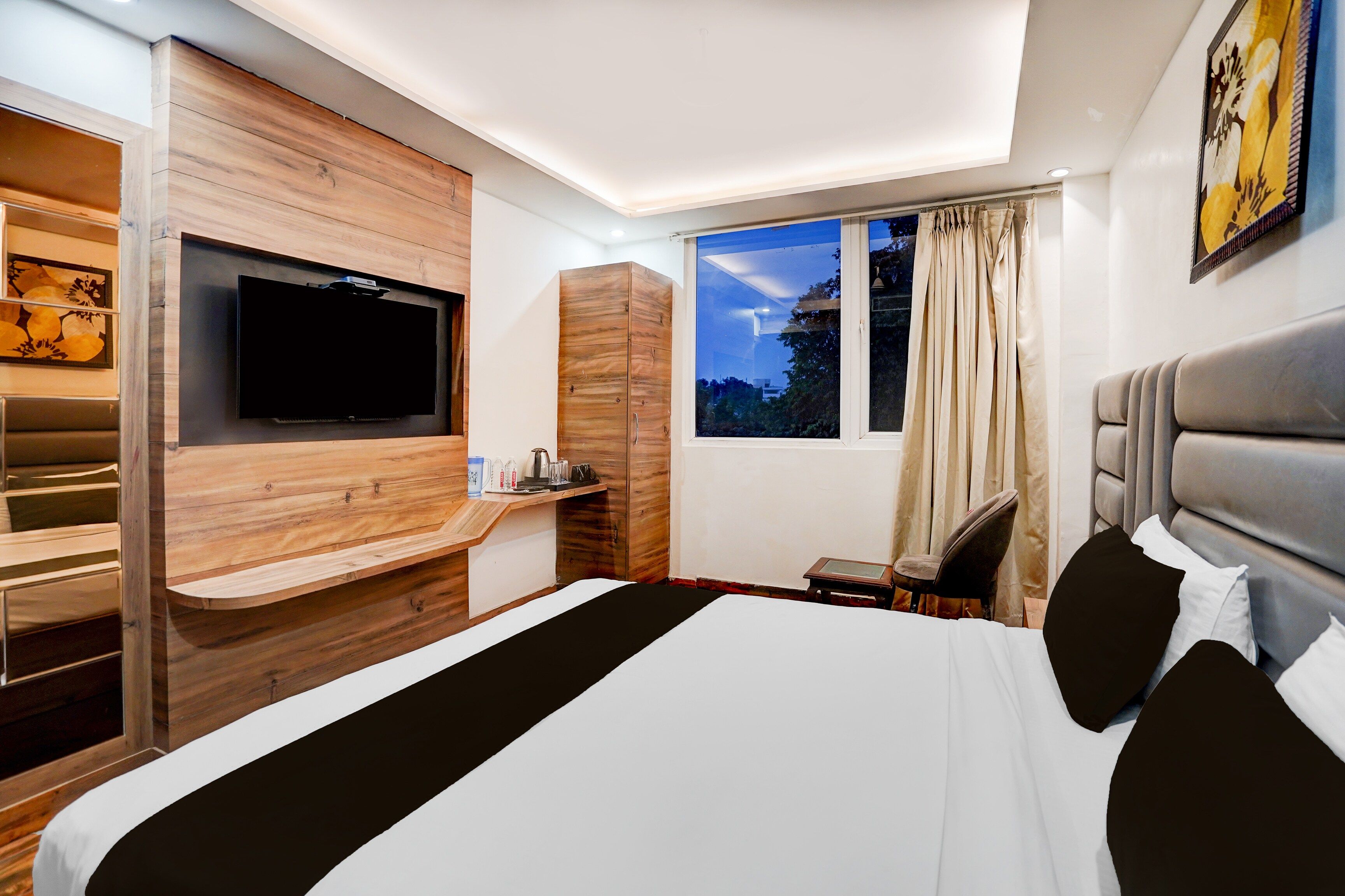 A modern hotel room with a large bed, a flat-screen TV mounted on a wooden wall, a small table with a kettle, and a window with curtains.