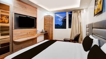 A modern hotel room with a large bed, a flat-screen TV mounted on a wooden wall, a small table with a kettle, and a window with curtains.