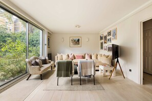 A living room with a sofa set, a chair, a television on a stand, and a large window with a view of greenery. - Kensington 2-Bed near Notting Hill with Garden (London)