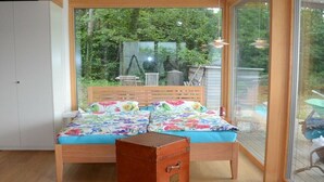 Une chambre avec un lit en bois, une valise et une grande fenêtre donnant sur un jardin.