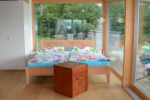 Ein Schlafzimmer mit einem hölzernen Bett, einem Koffer und einem großen Fenster mit Blick auf einen Garten.
