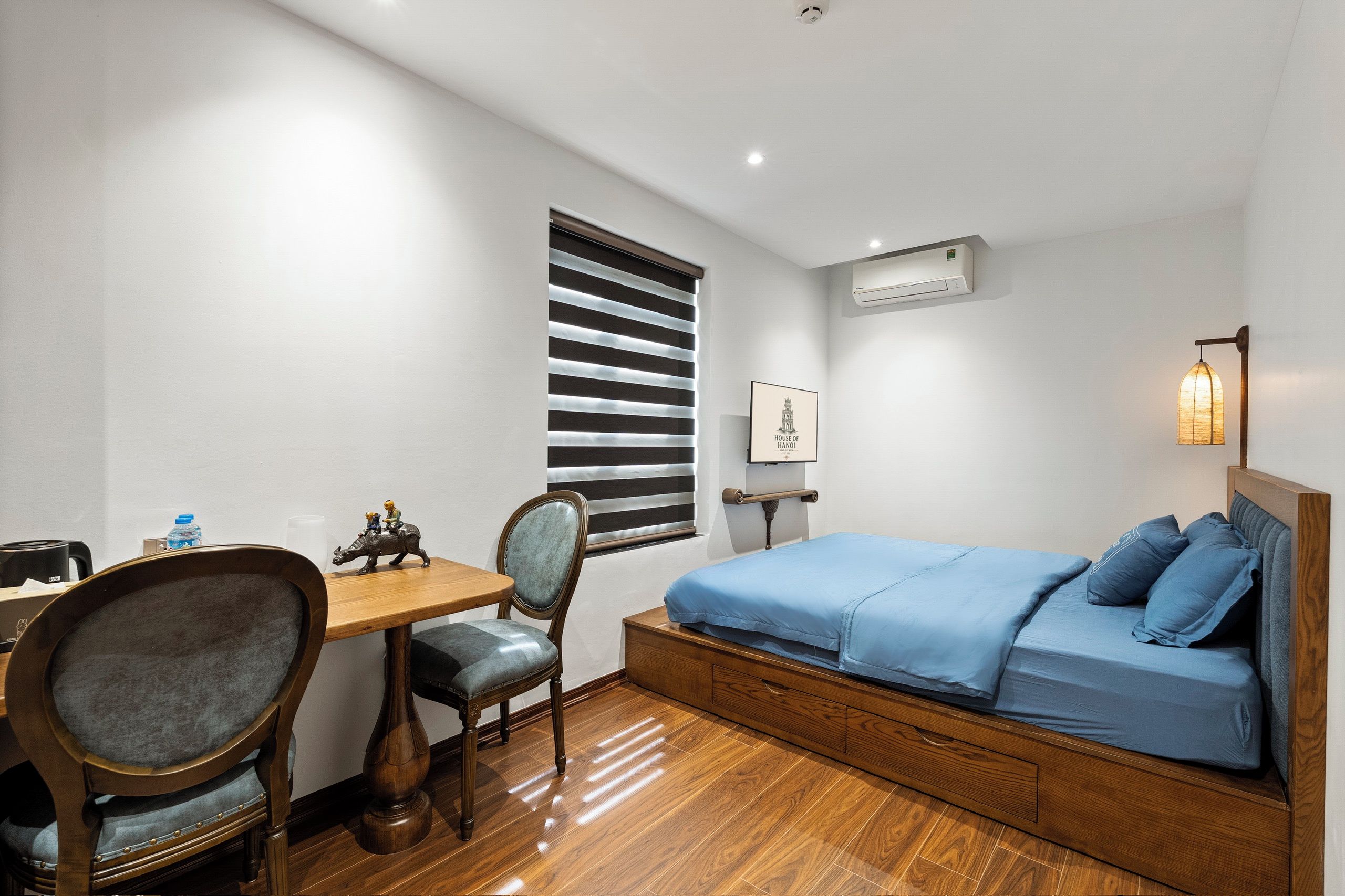 A bedroom with a bed, a desk, a chair, a window with blinds, an air conditioning unit, and a wall-mounted light fixture.
