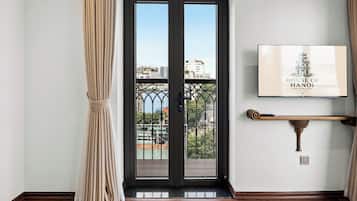 Entrada a una habitación de hotel con una puerta de vidrio de marco negro, cortinas color beige, piso de madera e imagen enmarcada de la Casa de Hanói en la pared.