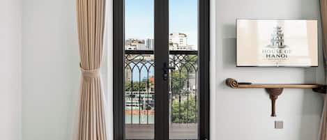 A hotel room entrance with a black-framed glass door, beige curtains, a wooden floor, and a framed picture of the House of Hanoi on the wall.