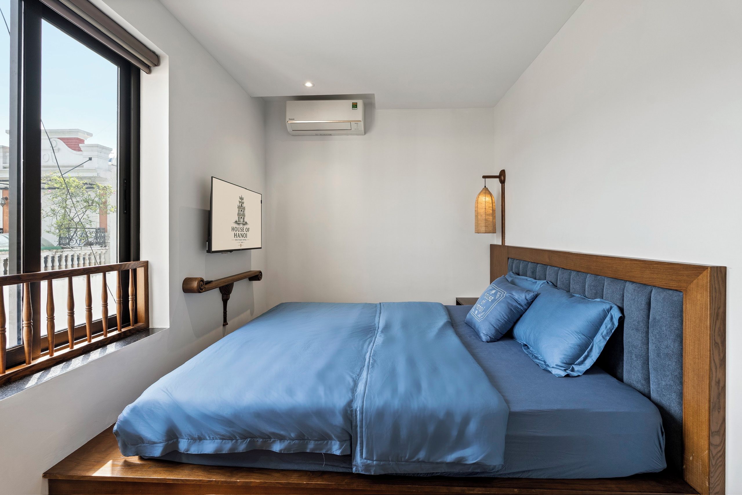 A hotel room with a bed, a window, an air conditioning unit, a framed picture, and a hanging light fixture.