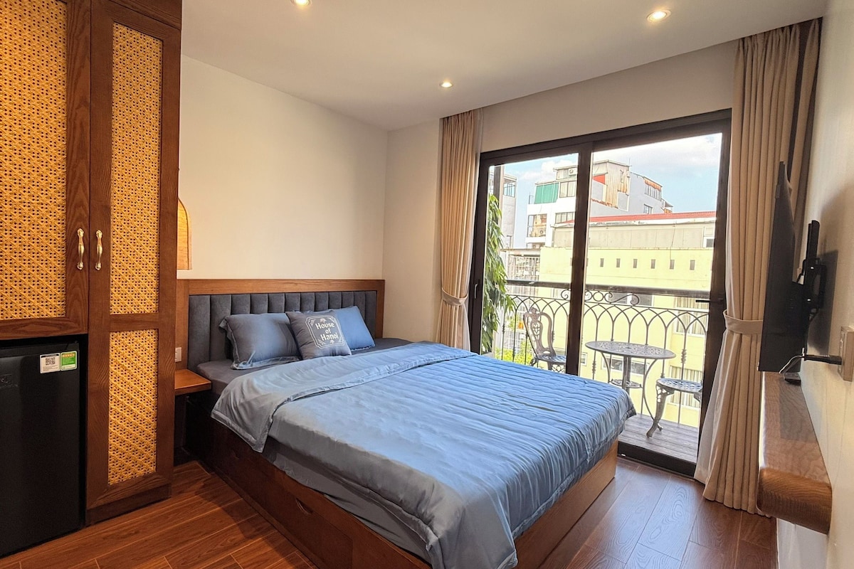 A bedroom with a bed, a wardrobe, a small fridge, and a balcony with a view of buildings.