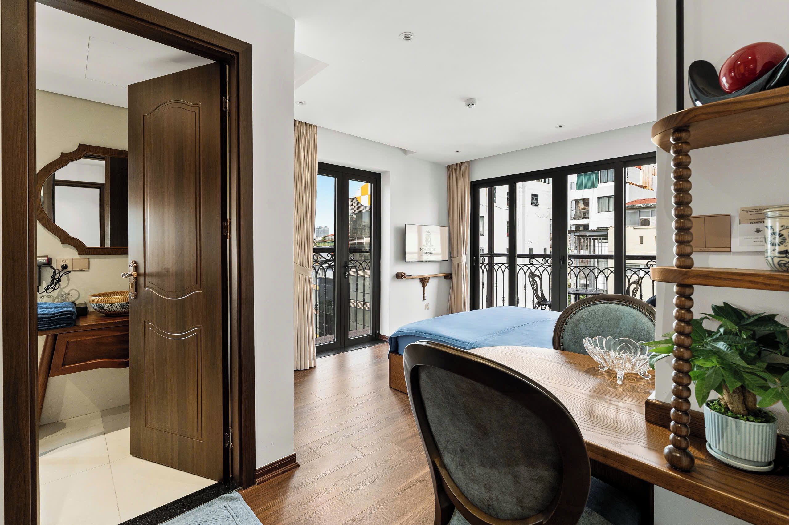 A hotel room with a wooden door, a bed with a blue bedspread, a dining table with chairs, a potted plant, and a view of the city through large windows.