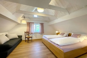 Quarto com teto estilo loft, cama com roupa de cama branca, sofá com almofadas, cadeira de madeira e janela com cortinas.