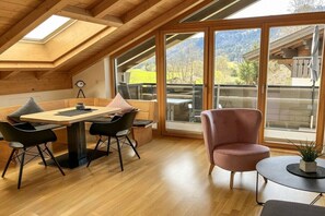 Quarto com teto de madeira, mesa de jantar com cadeiras, poltrona rosa e grandes janelas com vista para o verde.