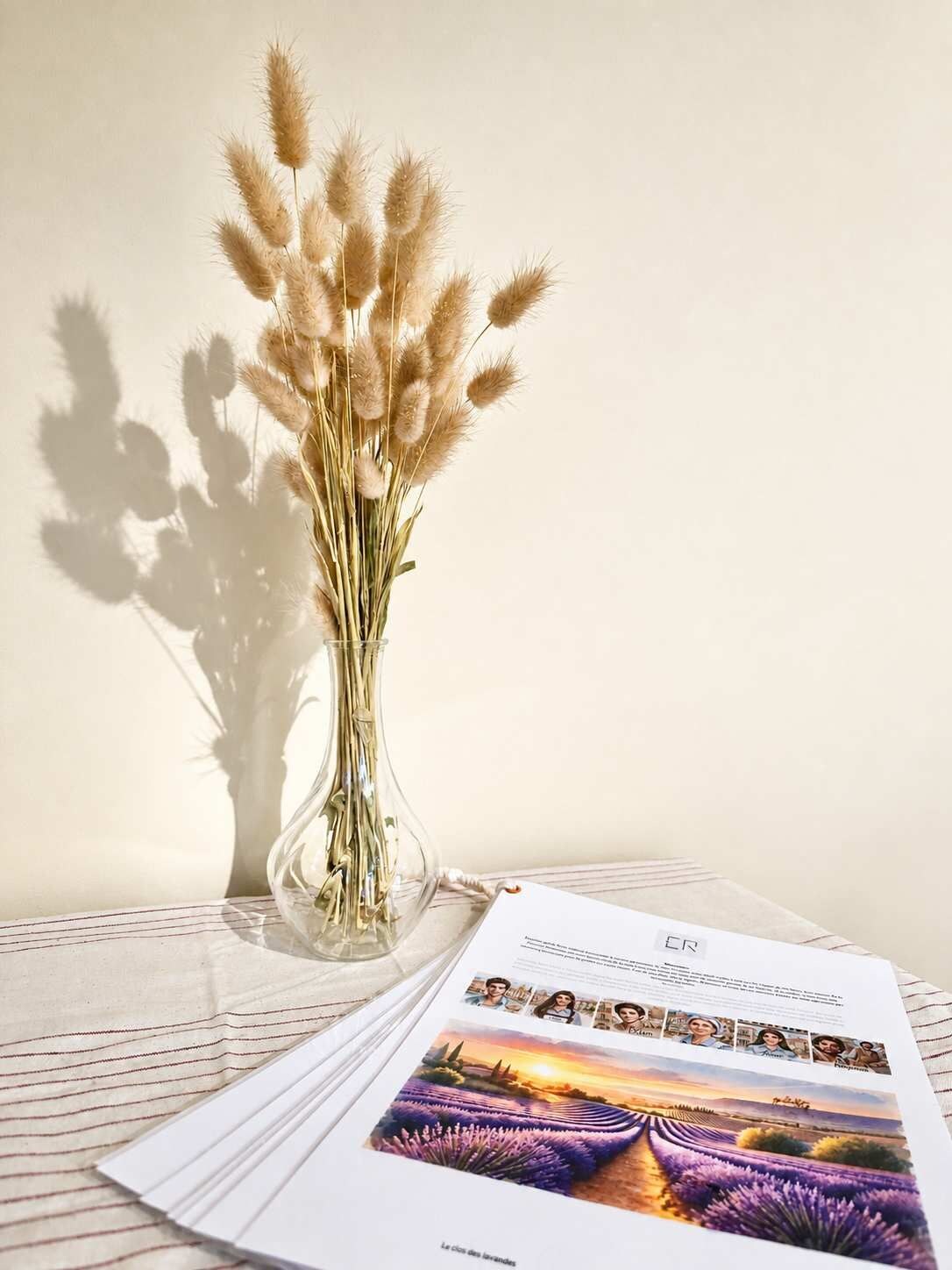 Un vase en verre transparent contenant des plumes de jarre séchées, placé devant un mur uni, un livre illustré avec un champ de lavande, et un tissu à rayures.