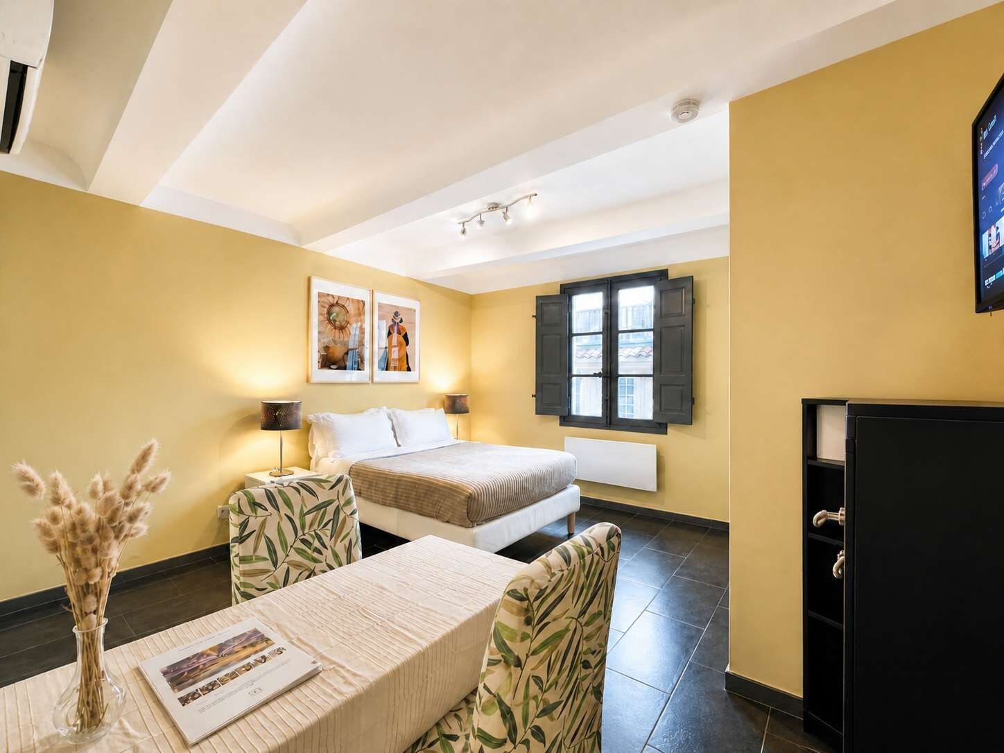 Habitación de hotel con cama, escritorio, dos sillas, un florero con flores secas y un cuadro en la pared.