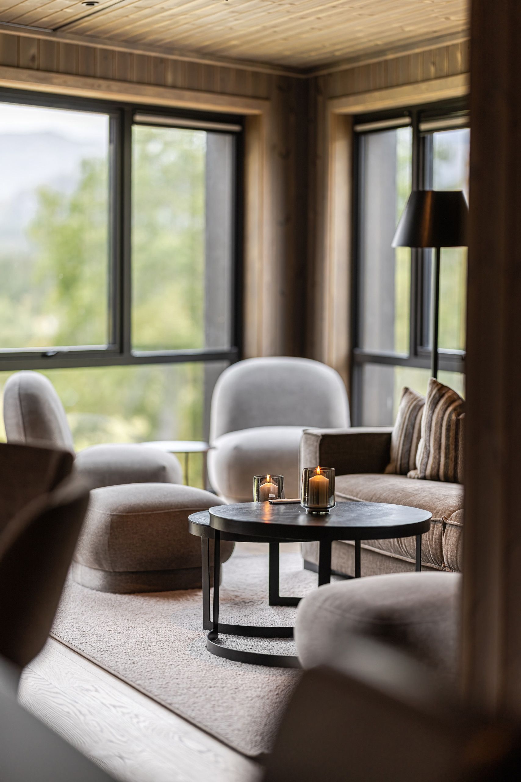 Ein modernes Wohnzimmer mit einer Couch, Sesseln und einem Couchtisch. Zwei Fenster bieten Blick auf die grüne Umgebung.