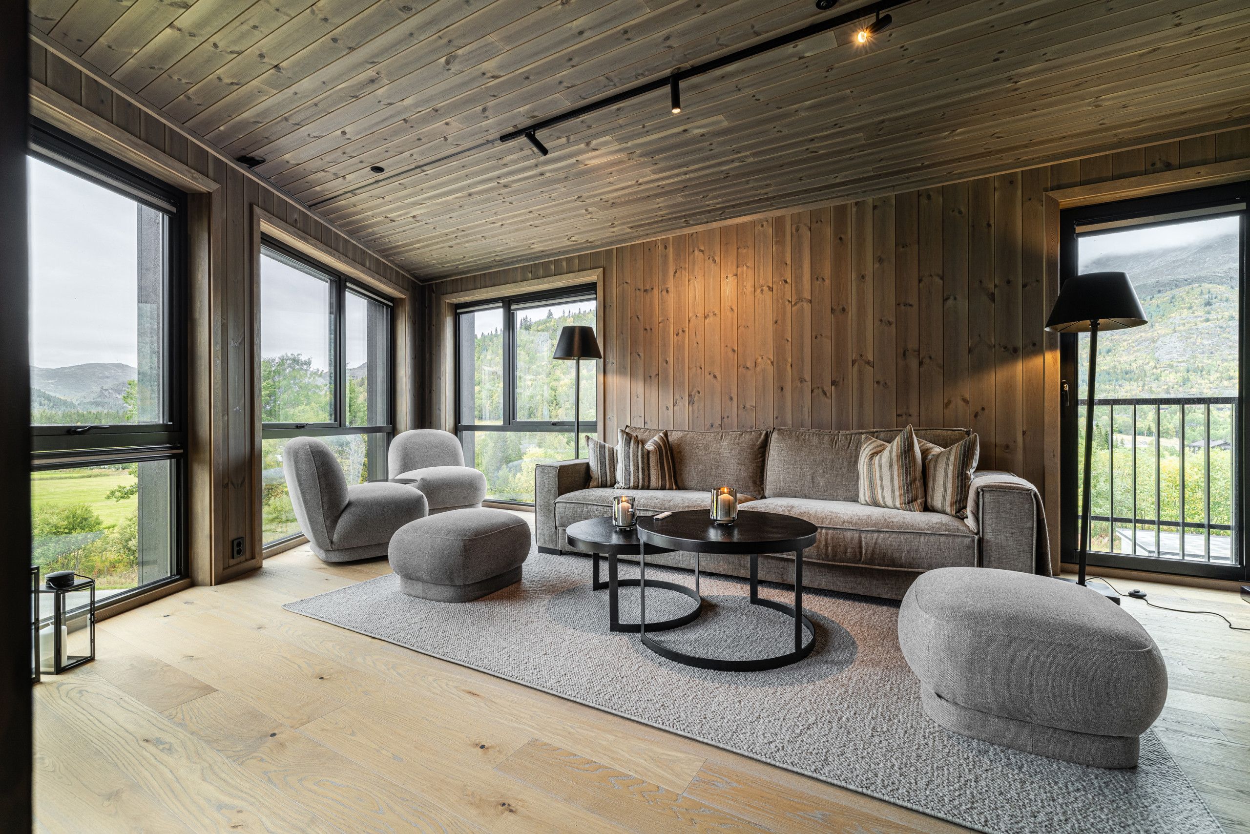 Amplia habitación con piso de madera, un sofá, sillones, una mesa de centro y ventanas amplias que ofrecen vistas al exterior.