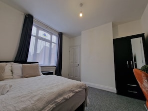 A bedroom with a bed, a window with curtains, a door, and a small table.
