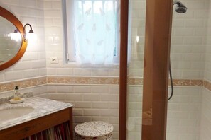 Ein Badezimmer mit weiß gefliesten Wänden, einem hölzernen Waschtisch mit Spiegel, einem kleinen runden Hocker und einer Duschkabine mit Glastür.