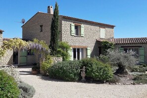 A stone house with a gravel driveway, green shutters, and a garden with various plants and a statue.