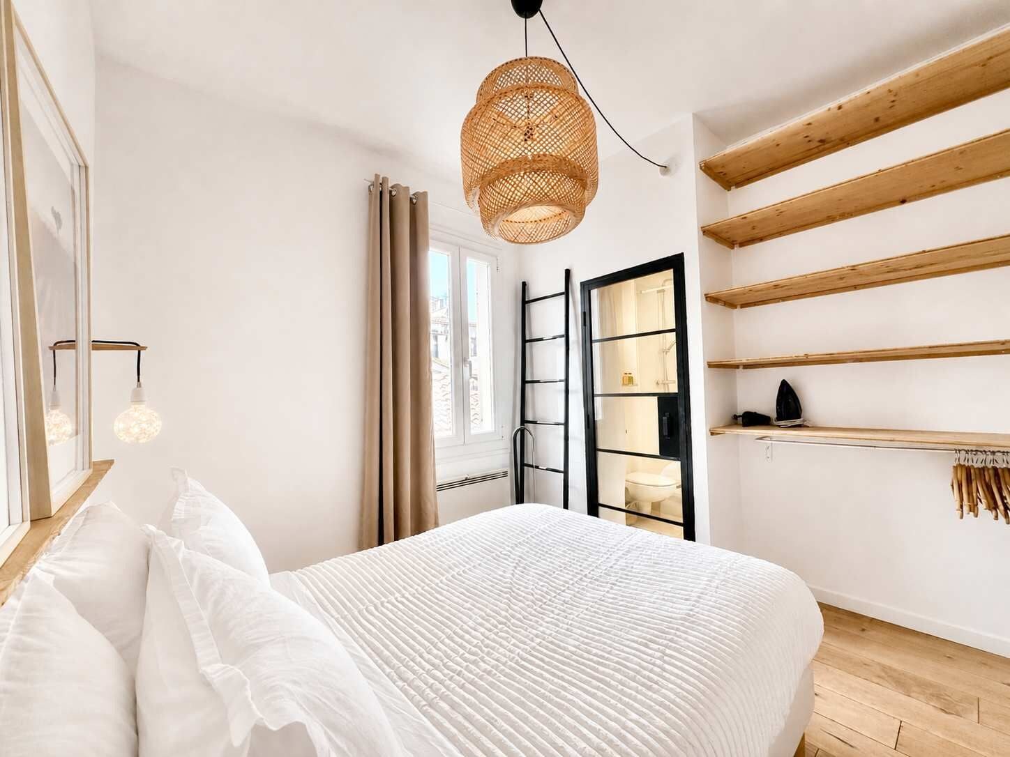 A bedroom with a bed, a hanging light fixture, a window with curtains, a radiator, and wooden shelves.