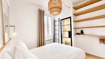 A bedroom with a bed, a hanging light fixture, a window with curtains, a radiator, and wooden shelves.