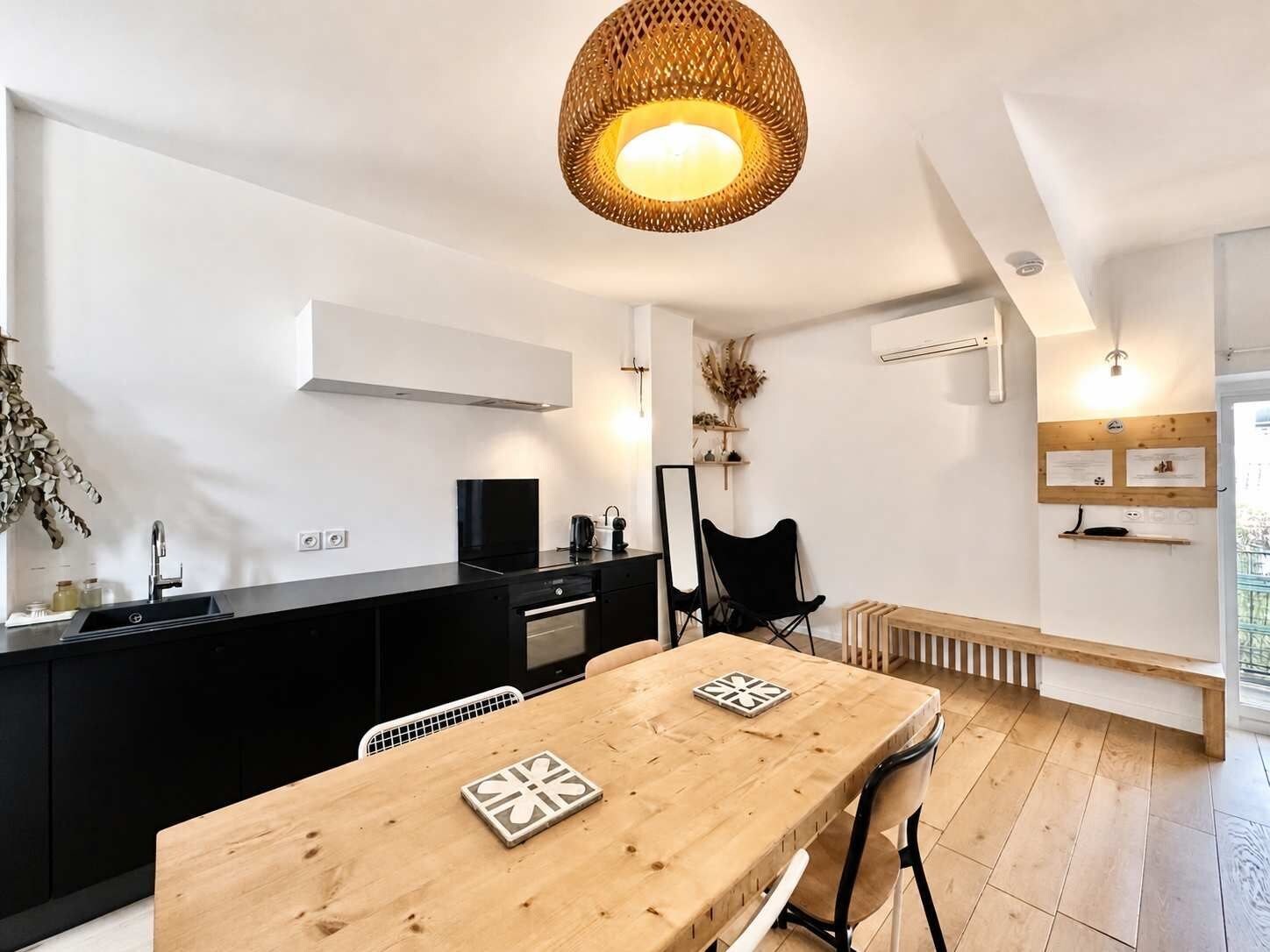 Une cuisine moderne avec un plan de travail noir, une table à manger en bois avec deux chaises, et un luminaire suspendu en fibres tressées.